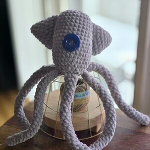 Op de afbeelding: Een handgemaakte, lavendelkleurige gehaakte octopus knuffel met een groot blauw knoopoog. De octopus heeft acht lange, krullende tentakels en wordt tentoongesteld op een gouden draadstandaard. Het speelgoed is gemaakt van zacht, getextureerd garen.