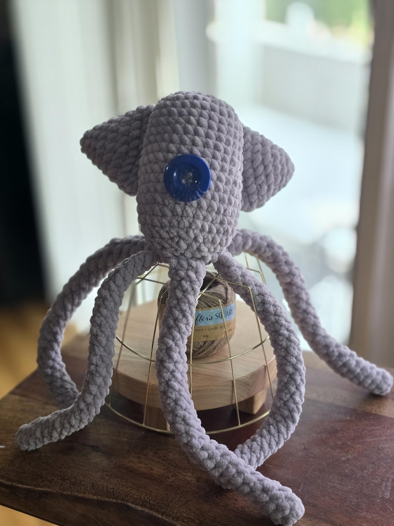 Op de afbeelding: Een handgemaakte, lavendelkleurige gehaakte octopus knuffel met een groot blauw knoopoog. De octopus heeft acht lange, krullende tentakels en wordt tentoongesteld op een gouden draadstandaard. Het speelgoed is gemaakt van zacht, getextureerd garen.