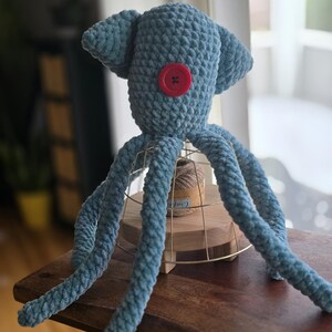 Op de afbeelding: Handgemaakte blauwe gehaakte octopuspop met een rode knoop als oog. Het pluche speelgoed heeft acht lange tentakels en twee oorachtige aanhangsels op zijn hoofd. De octopus staat op een houten oppervlak.