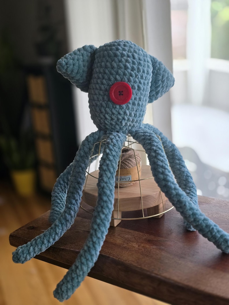 Op de afbeelding: Handgemaakte blauwe gehaakte octopuspop met een rode knoop als oog. Het pluche speelgoed heeft acht lange tentakels en twee oorachtige aanhangsels op zijn hoofd. De octopus staat op een houten oppervlak.