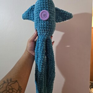 Op de afbeelding: Een teal gehaakte octopus knuffel met een grote paarse knoop en kleine vinnen. De knuffel heeft lange, bungelende tentakels. Het speelgoed is gemaakt van een zacht, getextureerd garen.