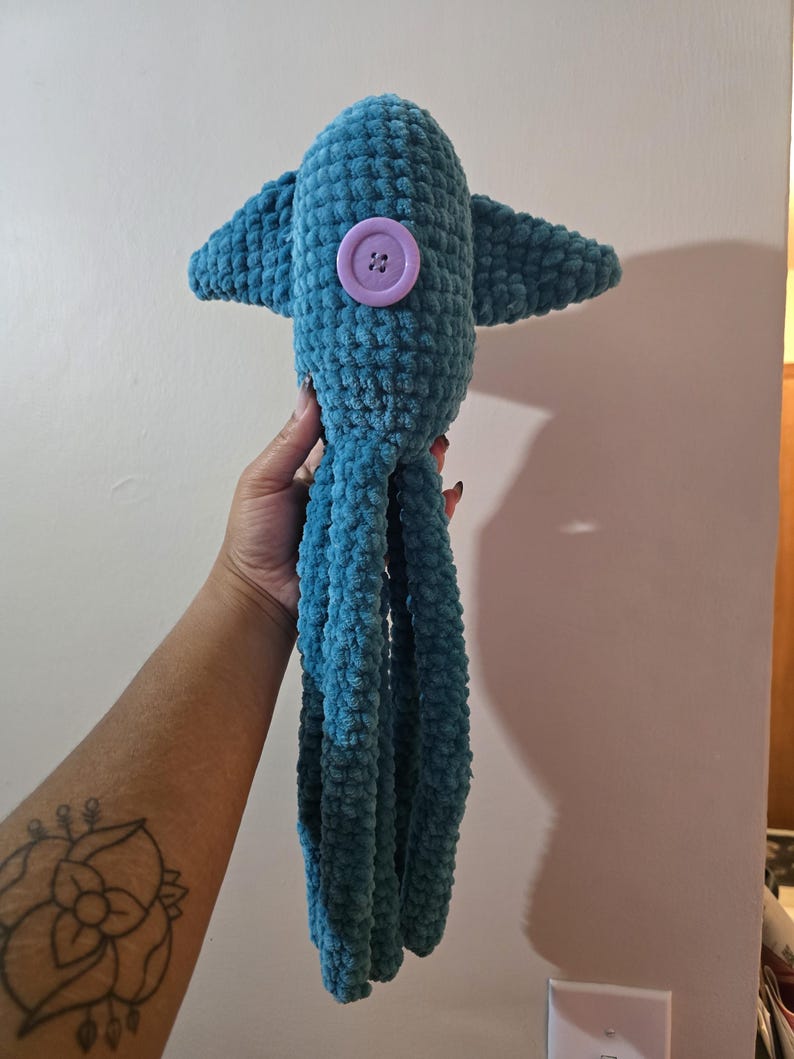 Op de afbeelding: Een teal gehaakte octopus knuffel met een grote paarse knoop en kleine vinnen. De knuffel heeft lange, bungelende tentakels. Het speelgoed is gemaakt van een zacht, getextureerd garen.