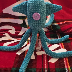 Op de afbeelding: Een handgemaakt turquoise gehaakt wezen met een afgeronde kop, twee zijvinnen en meerdere lange tentakels. Een lichtpaarse knoop is op de voorkant genaaid. Het pluche speelgoed wordt getoond op een rood-groene geruite deken.