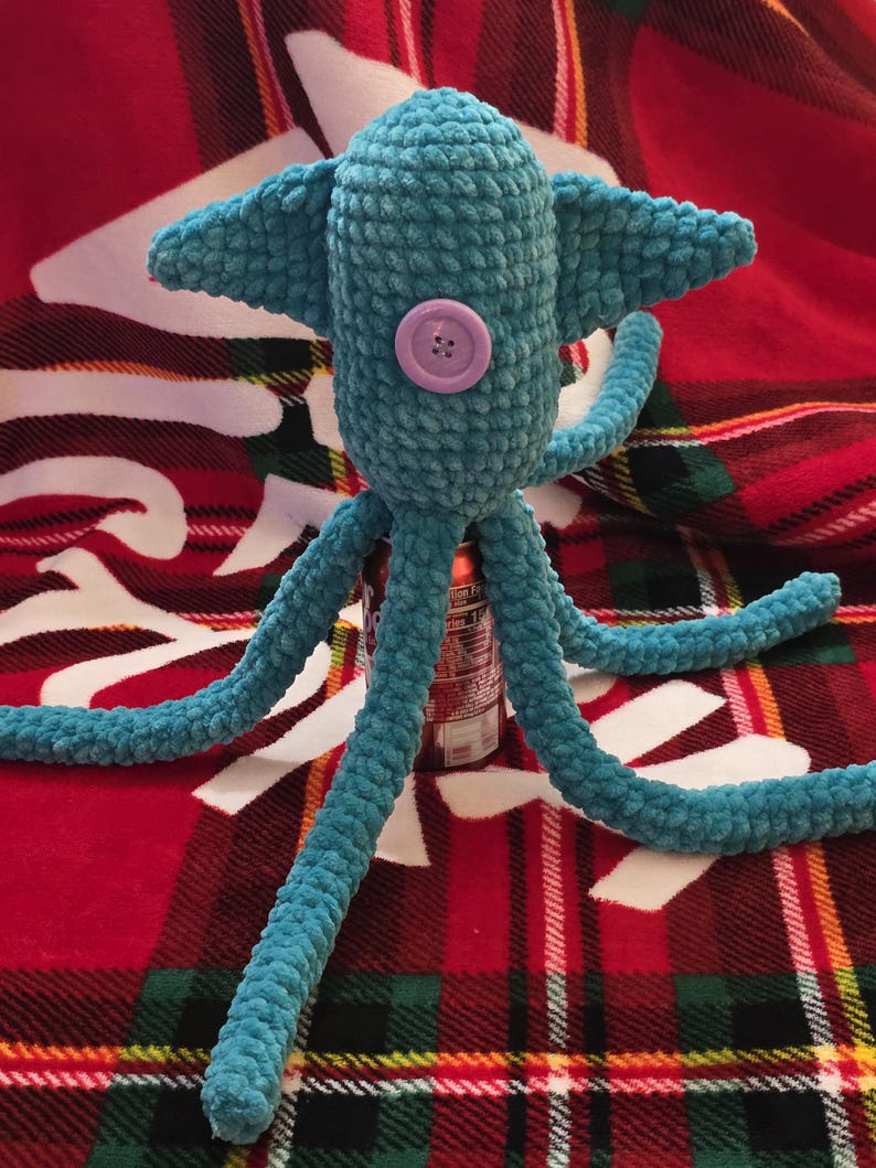 Op de afbeelding: Een handgemaakt turquoise gehaakt wezen met een afgeronde kop, twee zijvinnen en meerdere lange tentakels. Een lichtpaarse knoop is op de voorkant genaaid. Het pluche speelgoed wordt getoond op een rood-groene geruite deken.