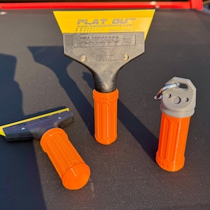 Puede incluir: Se muestran tres herramientas de detailing automotriz. Una espátula amarilla y negra con el texto "FLAT OUT" y "FUSION SHORTY 5" está en el centro. También se muestran dos herramientas adicionales con mangos naranjas. Una tiene una hoja amarilla y la otra una tapa gris.