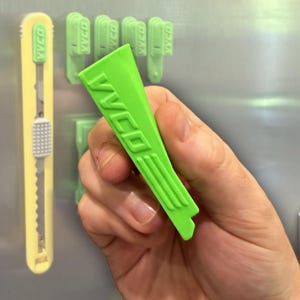 May include: A bright green plastic tool with the text "VVCO" is held in a hand. A yellow utility knife and several green clips with the same text are visible in the background. The tool has a unique shape with a notched end.