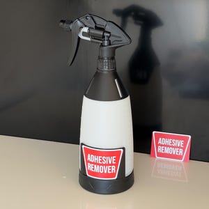 Puede incluir: Un pulverizador en blanco y negro con una etiqueta roja y blanca que dice "ADHESIVE REMOVER". La botella tiene un gatillo y una boquilla negros. Hay una segunda etiqueta al lado de la botella.