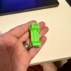 May include: A bright green 3D printed plastic object with a silver metal piece on top. The object has a rectangular shape with a small hole in the center.