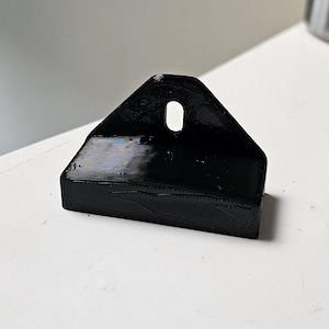 May include: A black 3D-printed mounting bracket with a rectangular base and a triangular top section featuring a single centered vertical oval hole for screw attachment. The surface shows visible layer lines from the additive manufacturing process.
