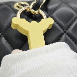 May include: A close-up of a yellow charm with a decorative design, attached to a gold-colored ring. The charm is set against a black quilted leather background, with a white fabric in the foreground.
