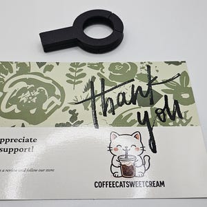 May include: A black coffee accessory sits above a thank you card. The card features a floral design, the words "Thank You", and a cartoon cat holding a coffee. The card also says "We appreciate your support!"