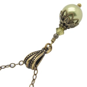 Pode incluir: Um close-up de um colar com um pingente de p&eacute;rola verde claro com uma tampa e detalhes decorativos em metal cor de bronze. O colar tem uma corrente e fecho de bronze, com uma pequena conta de cristal.