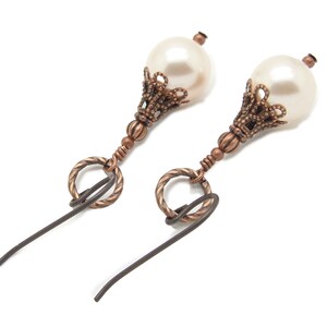 May include: A pair of earrings with a white pearl and a copper-colored filigree design. The earrings have a hook closure.