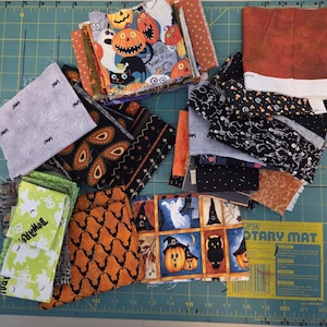 May include: A collection of assorted fabric squares featuring Halloween-themed patterns including pumpkins, ghosts, skeletons, bats, and spiders in orange, black, white, green, and purple colors, arranged in stacks on a green cutting mat.