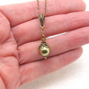 Pode incluir: Um colar de estilo vintage com corrente e pingente em tom de lat&atilde;o. O pingente apresenta uma grande conta redonda verde-oliva, acentuada com filigrana ornamental e contas menores. O colar &eacute; segurado na m&atilde;o de uma pessoa.