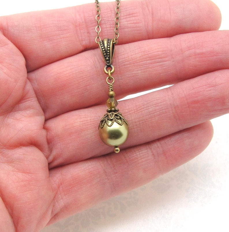 Pode incluir: Um colar de estilo vintage com corrente e pingente em tom de lat&atilde;o. O pingente apresenta uma grande conta redonda verde-oliva, acentuada com filigrana ornamental e contas menores. O colar &eacute; segurado na m&atilde;o de uma pessoa.