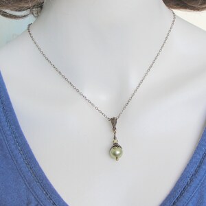Pode incluir: Um colar de estilo vintage com uma corrente delicada e um pingente com uma p&eacute;rola verde-claro. O pingente tem detalhes em metal ornamentados e pequenas contas transparentes. O colar &eacute; exibido em um manequim vestindo uma blusa azul.