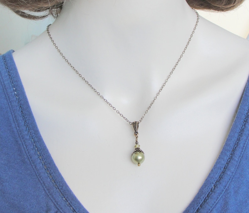 Pode incluir: Um colar de estilo vintage com uma corrente delicada e um pingente com uma p&eacute;rola verde-claro. O pingente tem detalhes em metal ornamentados e pequenas contas transparentes. O colar &eacute; exibido em um manequim vestindo uma blusa azul.