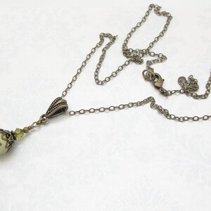 Pode incluir: Colar de estilo antigo com um pendente verde perolado. O pendente &eacute; adornado com pequenos cristais verde claro e uma tampa decorativa de tom bronze. A corrente e os elementos met&aacute;licos t&ecirc;m um acabamento bronze.