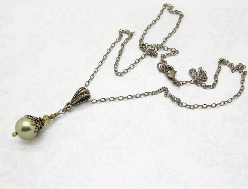 Pode incluir: Colar de estilo antigo com um pendente verde perolado. O pendente &eacute; adornado com pequenos cristais verde claro e uma tampa decorativa de tom bronze. A corrente e os elementos met&aacute;licos t&ecirc;m um acabamento bronze.