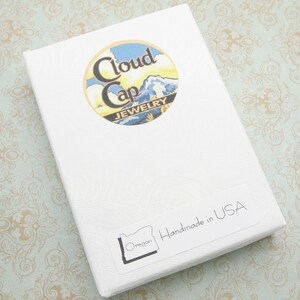 Pode incluir: Uma caixa de joias branca com uma superf&iacute;cie texturizada. A caixa apresenta um adesivo redondo com o texto "Cloud Cap Jewelry" e um design de paisagem. Um adesivo na caixa diz "Handmade in USA" e inclui o contorno do estado do Oregon.