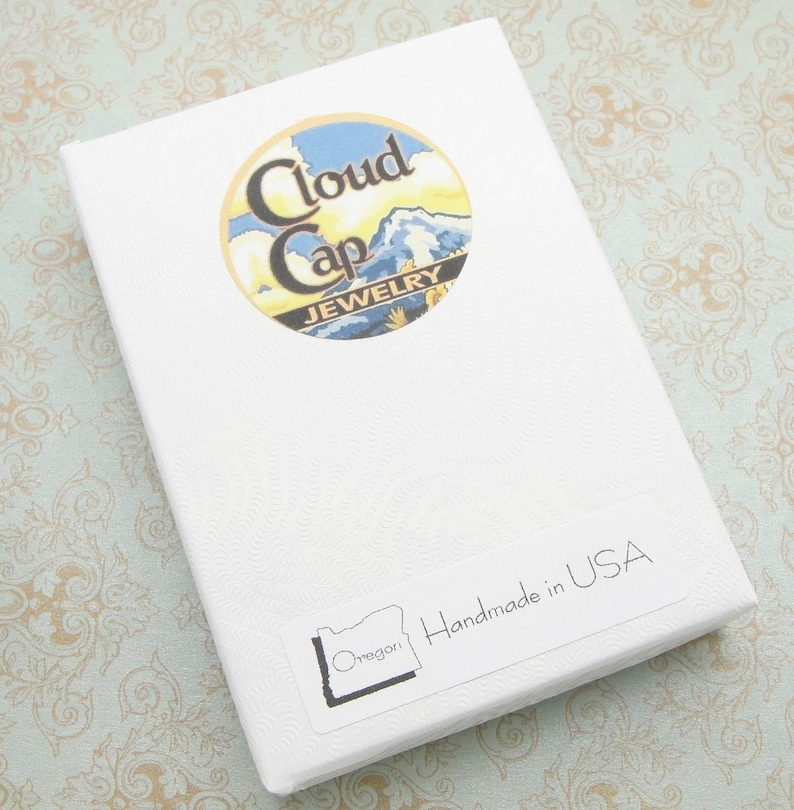 Pode incluir: Uma caixa de joias branca com uma superf&iacute;cie texturizada. A caixa apresenta um adesivo redondo com o texto "Cloud Cap Jewelry" e um design de paisagem. Um adesivo na caixa diz "Handmade in USA" e inclui o contorno do estado do Oregon.