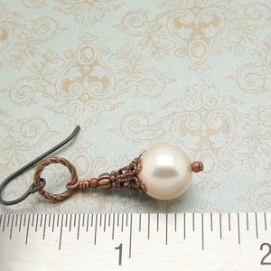 May include: A single copper-toned earring with a white pearl drop and a decorative filigree design. The earring is measured against a ruler.