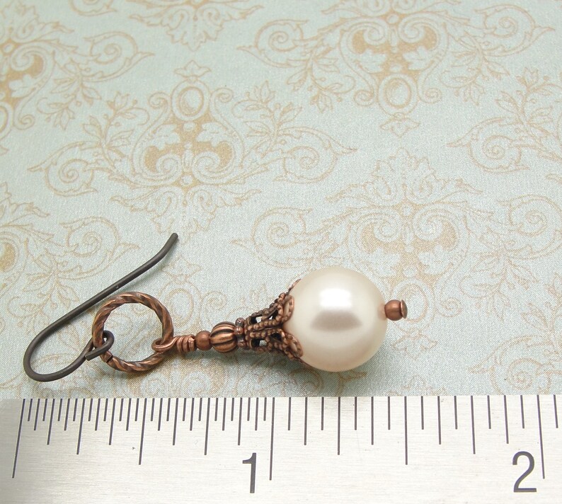 May include: A single copper-toned earring with a white pearl drop and a decorative filigree design. The earring is measured against a ruler.