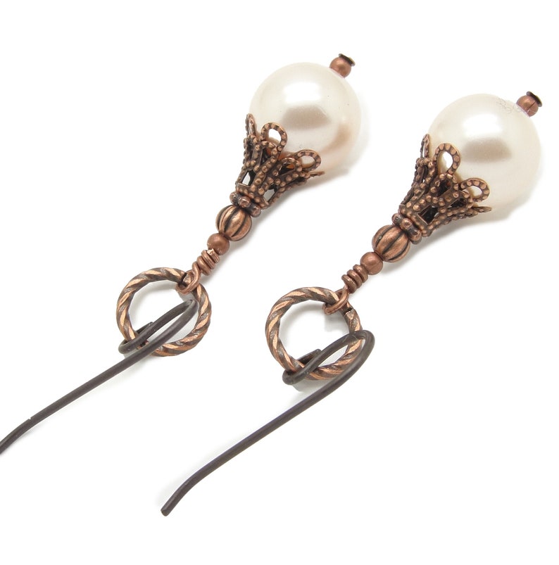 May include: A pair of earrings with a white pearl and a copper-colored filigree design. The earrings have a hook closure.