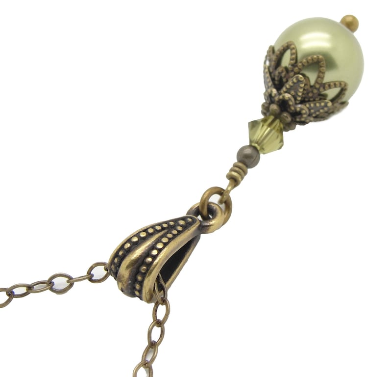 Pode incluir: Um close-up de um colar com um pingente de p&eacute;rola verde claro com uma tampa e detalhes decorativos em metal cor de bronze. O colar tem uma corrente e fecho de bronze, com uma pequena conta de cristal.