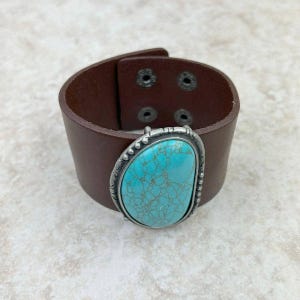 May include: A brown leather cuff bracelet with a large, oval turquoise stone set in a silver-colored metal frame. The bracelet has a buckle closure with four metal rivets.