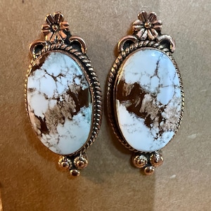 May include: A pair of oval-shaped earrings with white and brown marbled stones. The earrings have a copper-colored metal frame with a twisted rope design around the stone. A flower and three small beads adorn the top and bottom.