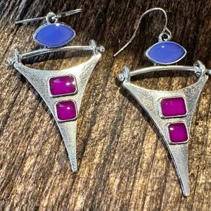 May include: A pair of silver-toned earrings with a geometric design. Each earring features a blue oval at the top, a silver bar, and a triangular shape with two magenta rectangular accents. The earrings are set against a wooden surface.