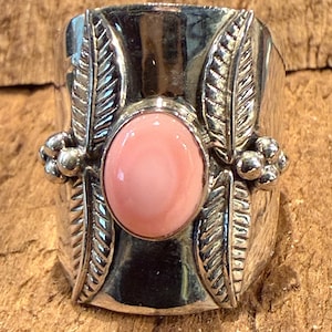 May include: A wide silver ring featuring a central oval-shaped smooth pink stone. The band is decorated with four embossed leaf motifs, two on each side of the stone, and small clusters of silver spheres positioned between the leaves on both sides.