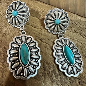 Könnte beinhalten: Silberfarbene Ohrringe mit türkisfarbenen Steinakzenten. Die Ohrringe haben oben ein Blumendesign und einen ovalen Anhänger mit einem türkisfarbenen Stein. Die Ohrringe sind im Südwest-Stil gestaltet.