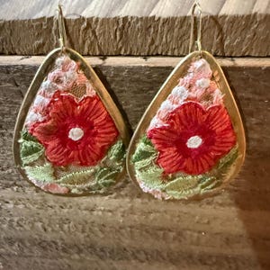 May include: A pair of teardrop-shaped earrings featuring gold-toned metal frames. The center of each earring is filled with white lace fabric embroidered with a vibrant red flower, a white center, and light green leaves at the bottom edge.