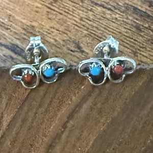 Puede incluir: Un par de pendientes de plata con cierre de tuerca. Cada pendiente presenta un pequeño diseño calado con una piedra turquesa central y una piedra roja. Los pendientes están colocados sobre una superficie de madera.