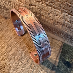 May include: A copper-toned cuff bracelet featuring a repeating embossed cross pattern along the center, flanked by textured vertical lines. The interior of the open-ended band displays three small circular magnets embedded on each side.