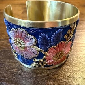 May include: A wide cuff bracelet featuring a polished gold-tone metal band and a deep blue fabric inlay. The fabric is decorated with embroidered pink flowers, blue leaf-like accents, and gold-tone metallic thread detailing with small sequins.