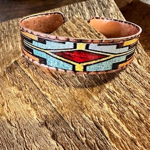 May include: Copper cuff bracelet with a geometric design featuring red, blue, yellow, and black accents. The bracelet has a rustic, open design and is displayed on a wooden surface. A unique piece of jewelry.