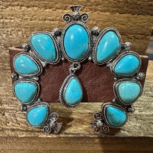 May include: A silver-toned metal cuff bracelet with turquoise-coloured stones. The bracelet features a horseshoe-shaped design with oval and teardrop-shaped turquoise stones. The bracelet is set on a brown leather band.