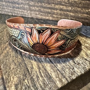 Pode incluir: Uma pulseira de couro bege com um design de girassol gravado à mão com pétalas laranja e amarelas, rodeada por intrincados arabescos pretos e verdes e um padrão de cestaria nas bordas com costuras visíveis.