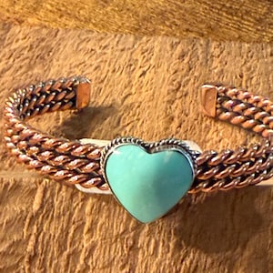 May include: A copper cuff bracelet featuring a triple-strand twisted wire design and a central heart-shaped turquoise stone set in a silver-toned bezel. The bracelet has an open back and a polished metallic finish with a smooth, light blue stone.