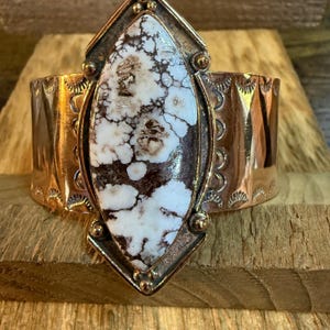 May include: A wide copper cuff bracelet with a large, oval white and brown stone centerpiece. The stone is set in a decorative frame. The cuff has etched patterns along the edges, creating a rustic, Southwestern style.