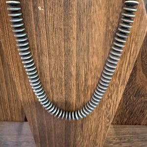 May include: A necklace featuring a long strand of circular, disc-shaped beads. The beads have a dark, matte center with metallic silver-toned edges, creating a uniform, repetitive pattern throughout the entire length of the jewelry piece.