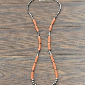 May include: A long necklace featuring alternating sections of orange-red beads and silver-colored, round beads. The necklace has a silver-colored chain clasp. The beads are strung on a dark background.