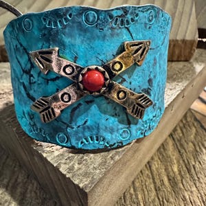 Puede incluir: Un brazalete turquesa con un acabado envejecido. El brazalete presenta flechas cruzadas en tonos bronce y oro, centradas con una piedra roja. El brazalete descansa sobre una superficie de madera, mostrando su encanto rústico.