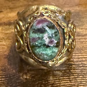 May include: A vintage-style ring with a large oval gemstone. The stone features a mix of green and maroon colors. The ring has a gold-colored metal band with ornate floral and scrollwork details.