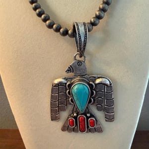 May include: A silver-toned metal necklace featuring a bird-shaped pendant with a central teardrop-shaped turquoise stone and three small rectangular red stones at the base. The pendant hangs from a strand of round, dark metallic beads.