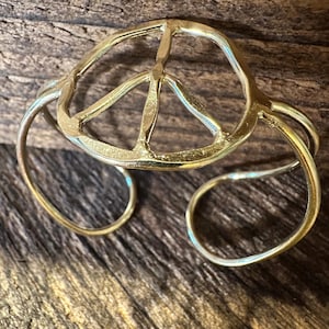 May include: A gold-colored cuff bracelet featuring a peace symbol design. The open-ended bracelet has a circular band that wraps around the wrist. The peace symbol is in the center of the bracelet.